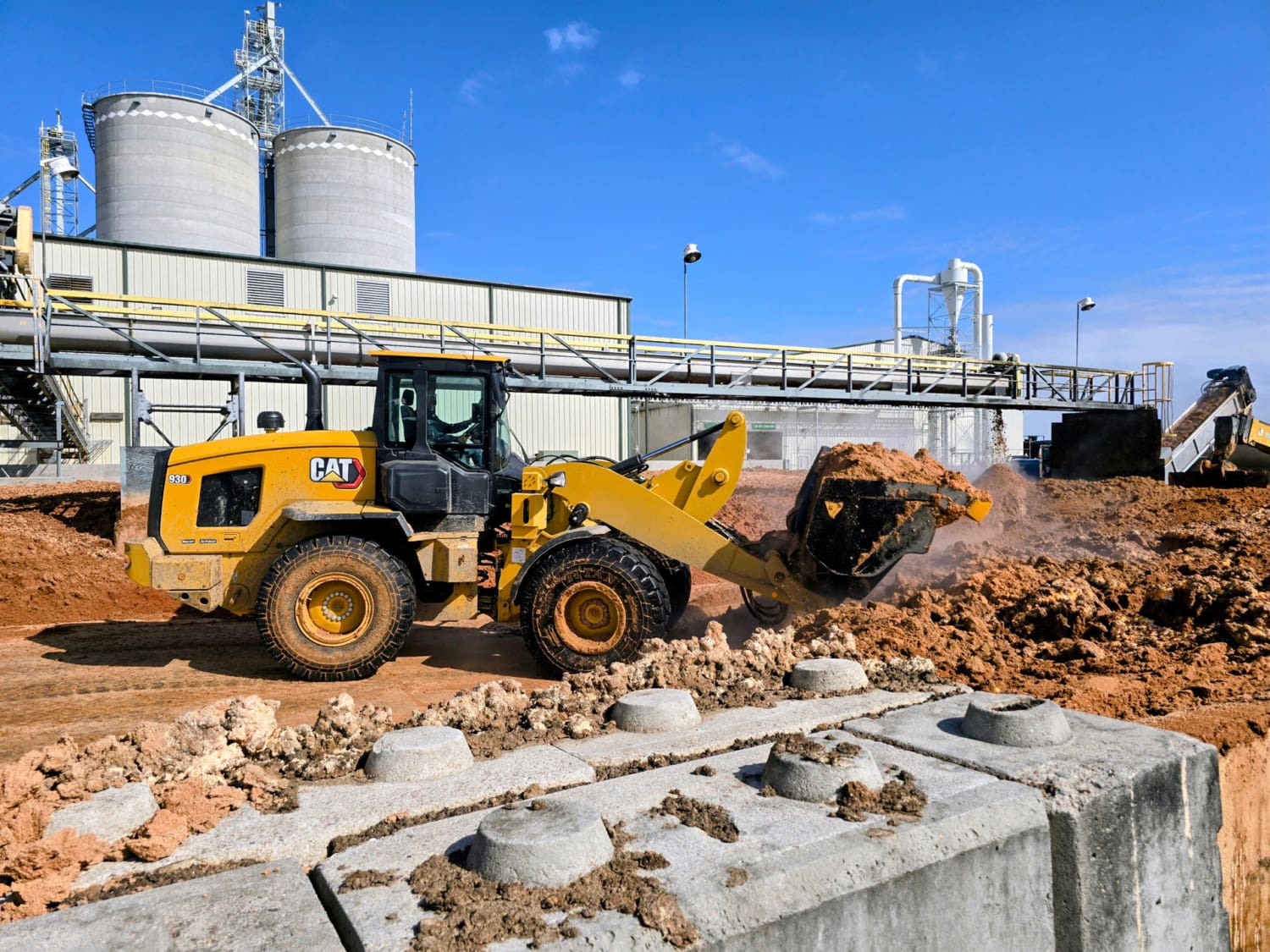 A CAT 930 high lift loader moves abrasive WDG at an ethanol processing plant, showing the heavy-duty Dymax bucket in action.